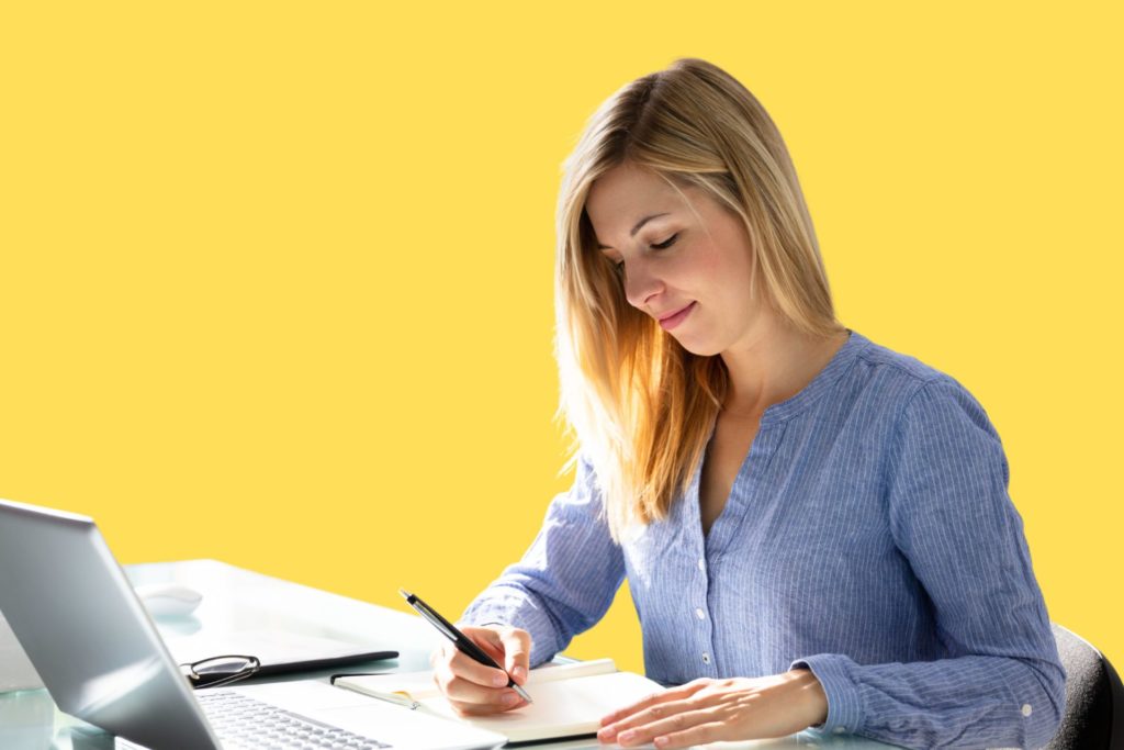 A woman writing on a paper, with a laptop in front of her.