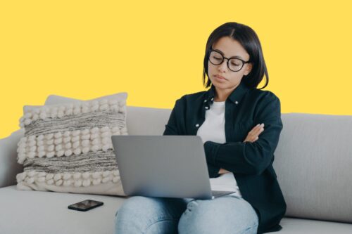 woman looking sceptical as she sits on a couch with her hands crossed, looking into her laptop