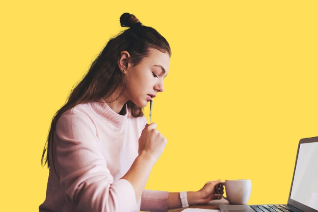 a woman holding a mug and sitting in front of a laptop, with a pen to her chin.