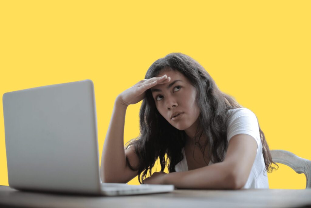 A person looking devastated with their hand on their forehead. With an open laptop in front of them on the desk.