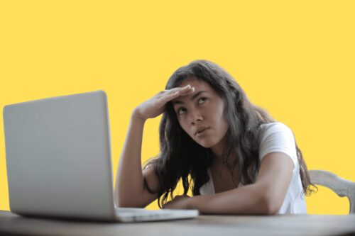 A person looking devastated with their hand on their forehead. With an open laptop in front of them on the desk.